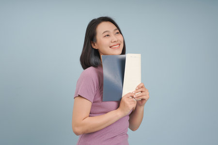 A cheerful Asian woman stands against a soft blue backdrop, holding two notebooks close to her...