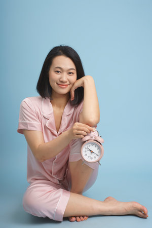 A young woman dressed in light pink pajamas sits comfortably on the floor, smiling gently while holding an alarm clock.の写真素材