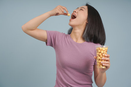 A cheerful young woman with short hair samples crispy popcorn from a container, expressing delight. She stands in front of a blue backdrop, radiating joy.の写真素材