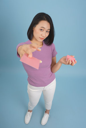 A woman poses confidently while holding a pink card in one hand and decorative pink items in the other. She stands against a soft blue background, showcasing her outfit and accessories.の写真素材
