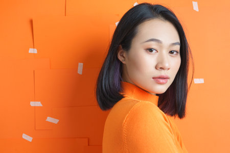 A woman with straight black hair wears a vibrant orange sweater, looking thoughtfully over her shoulder. The bright orange wall features playful strips of tape, enhancing the modern aesthetic.の写真素材