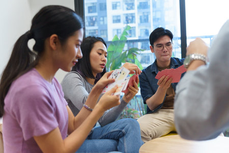 Four diverse friends are engaged in a lively card game in a bright indoor space filled with greenery. Laughter and excitement fill the air as they concentrate on their hands.の写真素材