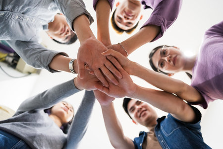 A group of Asian individuals stands together in a circle, placing their hands on top of each other...