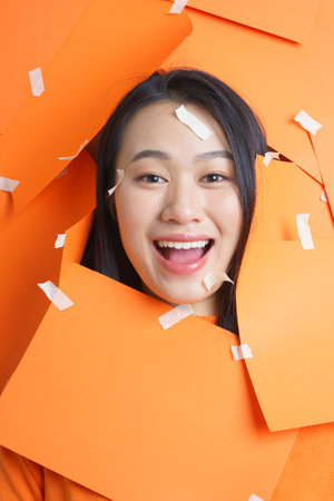 A young woman with long dark hair enjoys a playful moment as she is framed by vibrant orange paper....