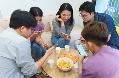 Friends gather around a low table filled with snacks, engaging with their smartphones. The atmosphere is relaxed, typical of a casual hangout in an Asian urban home.の写真素材