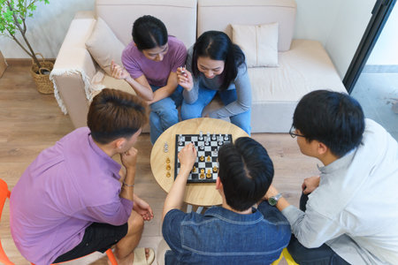 Five friends gather around a small table engaged in a chess game. The cozy indoor setting features a comfortable couch, natural light, and a relaxed atmosphere, creating a social vibe.の写真素材