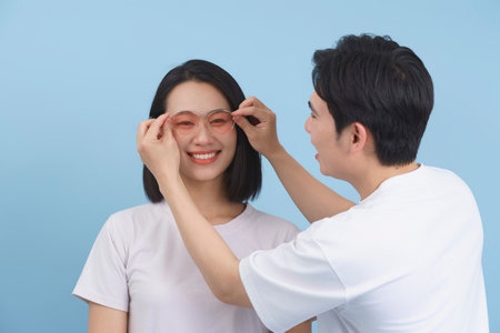 A man assists a woman in trying on trendy pink glasses, both smiling with joy in a light and airy blue studio. The atmosphere is playful and relaxed.の写真素材