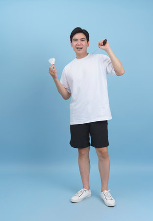A joyful young man stands in a light blue studio, smiling widely while holding a shuttlecock in one hand and a small racket in the other, exuding excitement and happiness.の写真素材
