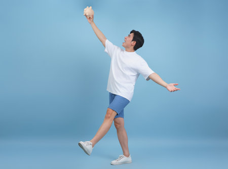 A young man with a cheerful expression stands confidently in a blue studio, joyfully raising a piggy bank above his head.の写真素材