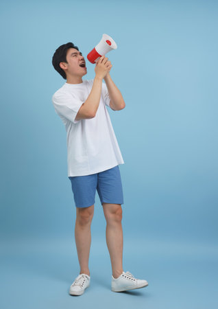 A young Asian man is energetically speaking into a red and white megaphone while standing against a solid blue background. His enthusiasm is palpable as he engages the audience.の写真素材