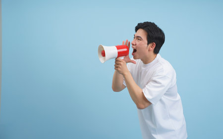 A young man passionately addresses a crowd using a megaphone. He is dressed in a plain white shirt and stands against a soft blue backdrop, conveying energy and engagement.の写真素材