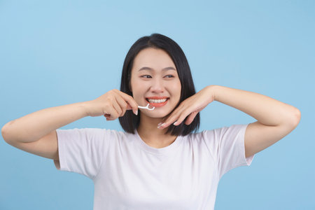 A young woman with straight black hair is happily flossing her teeth while smiling brightly. She is dressed in a simple white t-shirt and is focused on maintaining her dental hygiene.の写真素材