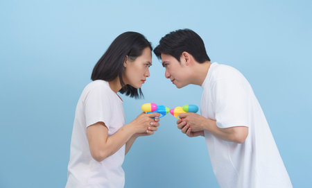 Two friends face each other, each holding bright, colorful water guns, showcasing expressions of playful competition. The soft blue background sets a lighthearted atmosphere.の写真素材