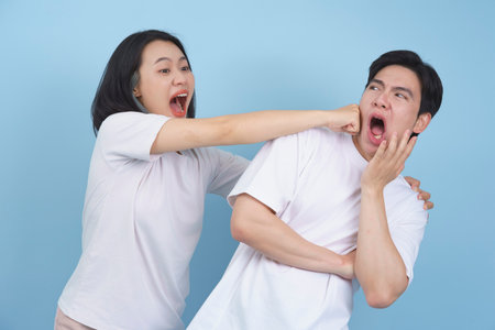 A woman is teasing a man by playfully pinching his cheek. Both are expressing exaggerated emotions, showcasing a light-hearted moment filled with laughter against a solid blue backdrop.の写真素材