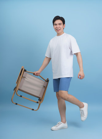 A young man with short hair smiles while lifting a wooden chair. He wears a white t-shirt and shorts, showcasing a lively and upbeat attitude in a light blue setting.の写真素材