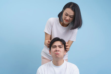 A woman smiles mischievously while styling a man's hair, who appears slightly uncomfortable. The...