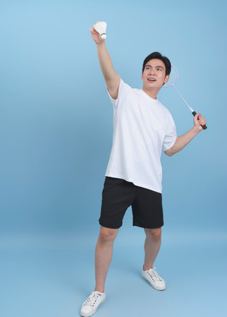Bright blue background features a young Asian man enthusiastically playing badminton. He is smiling while holding a racket and shuttlecock, showcasing his enjoyment during sports.の写真素材