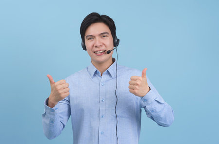 A young man wearing a light blue button-up shirt and a headset is smiling broadly while giving two thumbs up. The bright blue background enhances a cheerful atmosphere.の写真素材