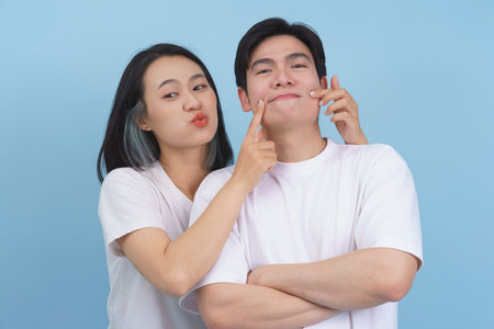 Two young adults enjoy a playful moment, with one person making a funny face while the other playfully pinches their cheek. They stand against a bright blue background, radiating joy.の写真素材