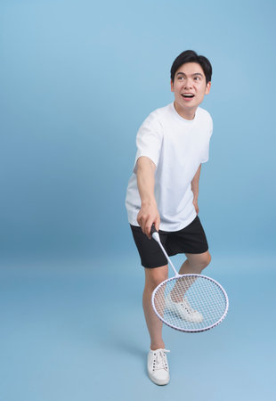 A young man with short dark hair stands in a bright blue studio, holding a badminton racket and looking excited. He is wearing a casual white t-shirt and black shorts, ready to play.の写真素材