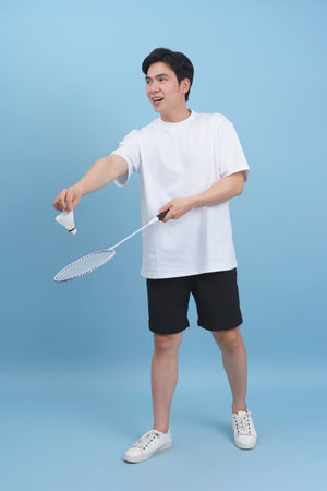 A young Asian man is engaged in a badminton match, holding a racket and preparing to hit a shuttlecock. He displays enthusiasm in a bright indoor setting with a blue background.の写真素材