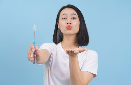 A cheerful young woman stands against a light blue background, playfully blowing a kiss while holding a toothbrush. Her joyful expression reflects a fun morning routine.の写真素材