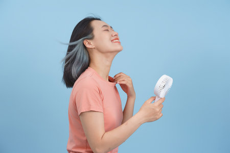 A young woman with black and blue hair smiles joyfully while holding a portable fan. She appears relaxed and cheerful, enjoying the cool breeze on a warm day with a soothing light blue backdrop.の写真素材