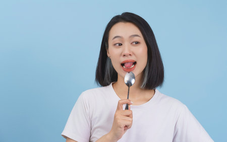 A young Asian woman is playfully holding a spoon with a dessert in her mouth, smiling brightly against a light blue backdrop. The atmosphere feels relaxed and joyful.の写真素材