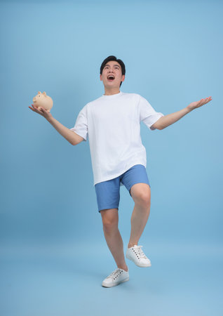A young man stands with one leg raised, displaying enthusiasm while holding a piggy bank. He is dressed in a white shirt and blue shorts, set against a soft blue backdrop.の写真素材