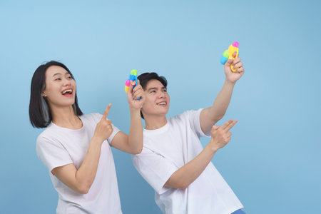 A young couple stands against a light blue backdrop, both smiling brightly. They point their colorful water guns playfully, exuding energy and enjoyment.の写真素材