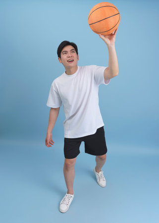 A young man is smiling while lifting a basketball above his head in a casual pose, dressed in a simple white shirt and black shorts. The bright blue backdrop creates a vibrant atmosphere.の写真素材