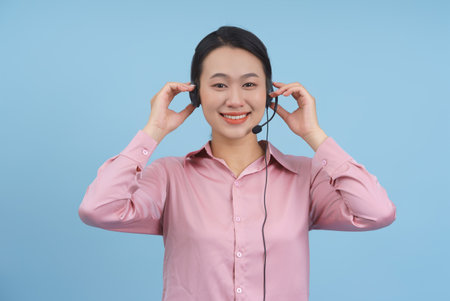 A smiling young woman adjusts her headset while standing in front of a solid blue background. She...