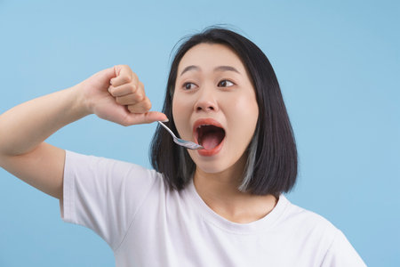 A smiling young woman takes a big bite with a spoon, showcasing enthusiasm for her meal. She is wearing a simple white shirt and has a playful demeanor.の写真素材