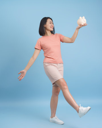 A cheerful young woman stands on one leg, holding a piggy bank high in her right hand.の写真素材