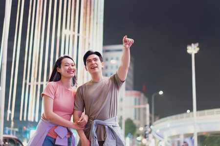 A couple stands close together in Hanoi, Vietnam, pointing and smiling as they admire the colorful lights showcasing the city's nightlife. Joy fills the air as they explore together.の写真素材