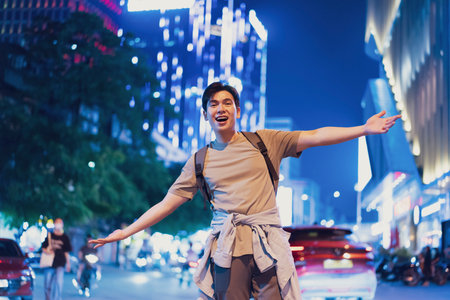 A young man stands confidently in the lively streets of Hanoi, Vietnam, expressing joy as he enjoys the vibrant nightlife. Colorful lights illuminate the urban scene around him.の写真素材