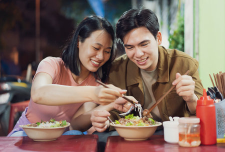 A young couple shares a joyful moment as they savor delicious Vietnamese dishes at a vibrant street stall in Hanoi. Their smiles reflect the warmth of the local dining experience at night.の写真素材