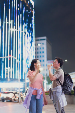 A couple takes a moment to savor ice cream while standing on a bustling street in Hanoi at night....