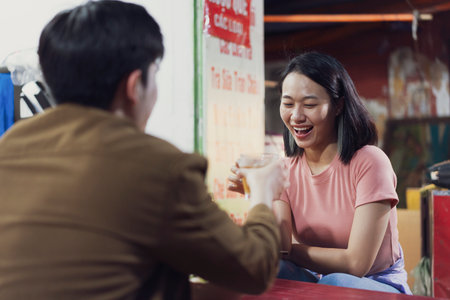 A young couple enjoys drinks together at a vibrant street stall in Hanoi, Vietnam. Laughter fills the air as they engage in friendly conversation, showcasing a relaxed evening atmosphere.の写真素材
