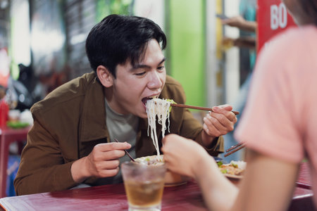 A young man is savoring a bowl of traditional Vietnamese noodles, using chopsticks to lift the noodles to his mouth. He enjoys this casual dining experience in Hanoi.の写真素材