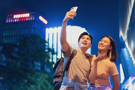 A young couple stands in the bustling streets of Hanoi at night, capturing a joyful selfie amidst the vibrant neon lights. The atmosphere is lively and fun, representing the city's youthful spirit.の写真素材