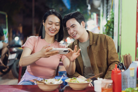 A young couple sits at a vibrant street food stall in Hanoi, Vietnam. They share a light moment...