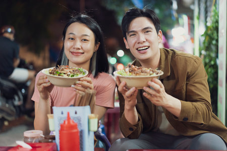 A young couple smiles while holding bowls of Vietnamese noodles at a street eatery in Hanoi. The...