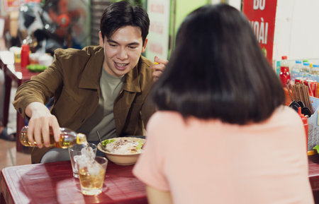 A man pours a drink while seated across from a woman at a table in a lively Hanoi eatery. They share...