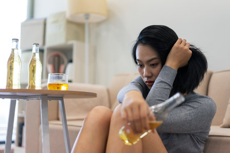 A young woman sits in a comfortable living room, looking pensive as she holds a drink. Glasses and...