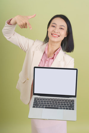 A young Asian woman is smiling and pointing at a blank laptop screen while wearing a professional outfit. The light green background adds a fresh, vibrant touch to her confident demeanor.の写真素材