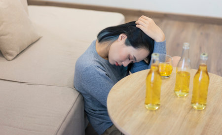 A young woman appears to be in deep thought while sitting on the floor next to a table. Empty bottles and a glass are scattered nearby in a cozy, modern room. Her posture suggests contemplation.の写真素材