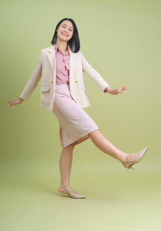A joyful Asian woman strikes a playful pose, her left leg raised slightly while wearing a light-colored business suit. Her smile adds to the vibrant atmosphere of the soft green background.の写真素材