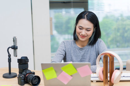 A young Asian woman sits at a desk in a stylish workspace, using a laptop and reviewing notes....
