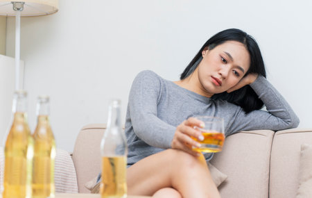 A woman wearing a gray sweater sits on a couch, holding a glass of drink and appearing thoughtful. She is surrounded by bottles, creating a cozy atmosphere in her living space during the evening.の写真素材